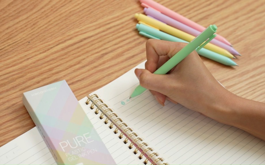 Hand writing in a spiral notebook with a mint gel pen, pastel pens and their box on a wooden desk